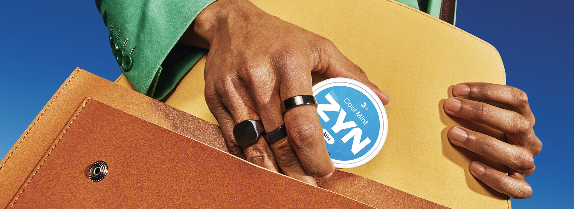 A person in a green suit placing a white ZYN nicotine pouches container inside an orange satchel behind a blue background.